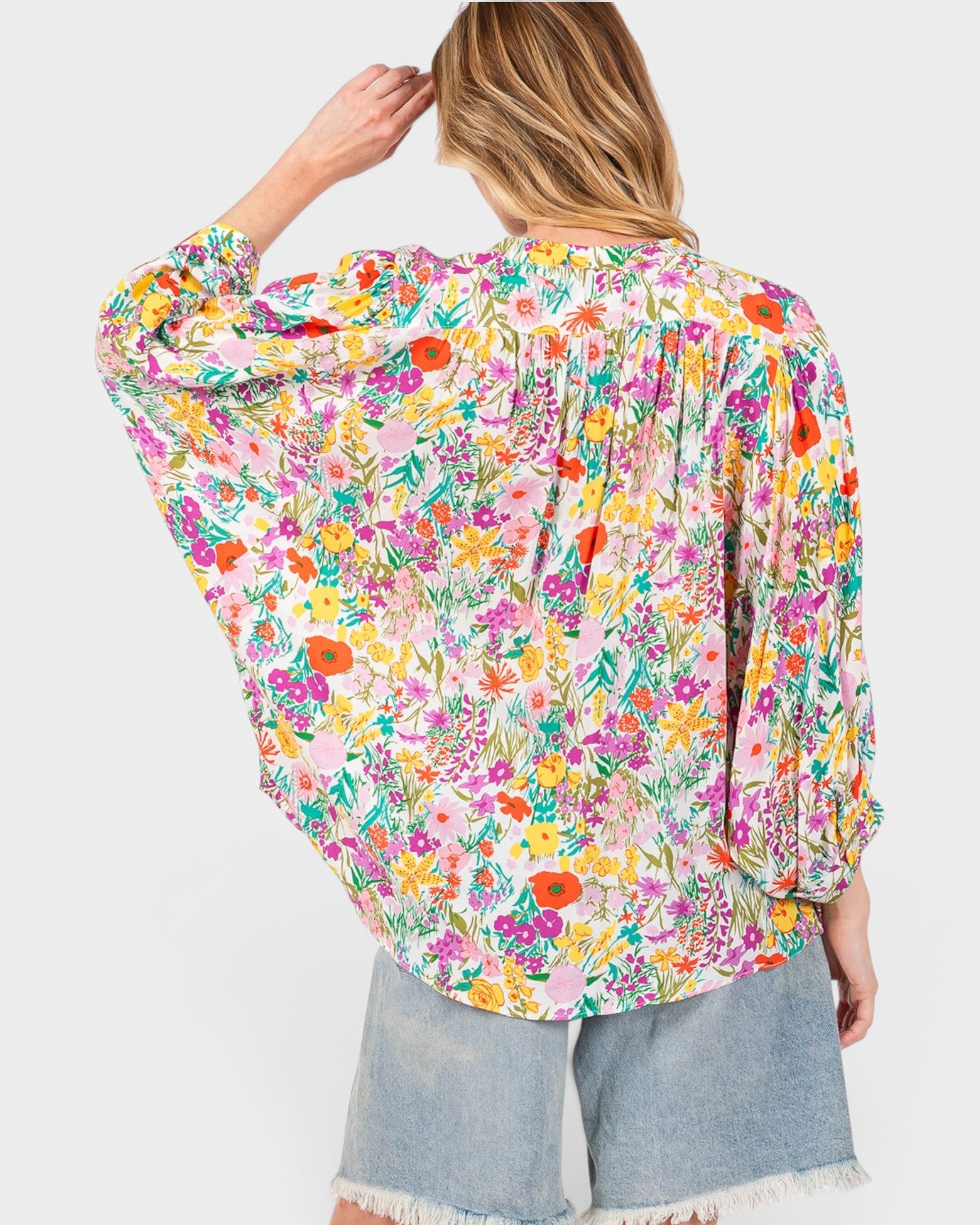 A woman shown from the back wearing a long-sleeve, loose-fitting blouse with a dense multicolor floral print on a light pink base, paired with light-wash denim shorts against a plain light gray background.