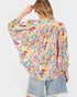 A woman shown from the back wearing a long-sleeve, loose-fitting blouse with a dense multicolor floral print on a light pink base, paired with light-wash denim shorts against a plain light gray background.