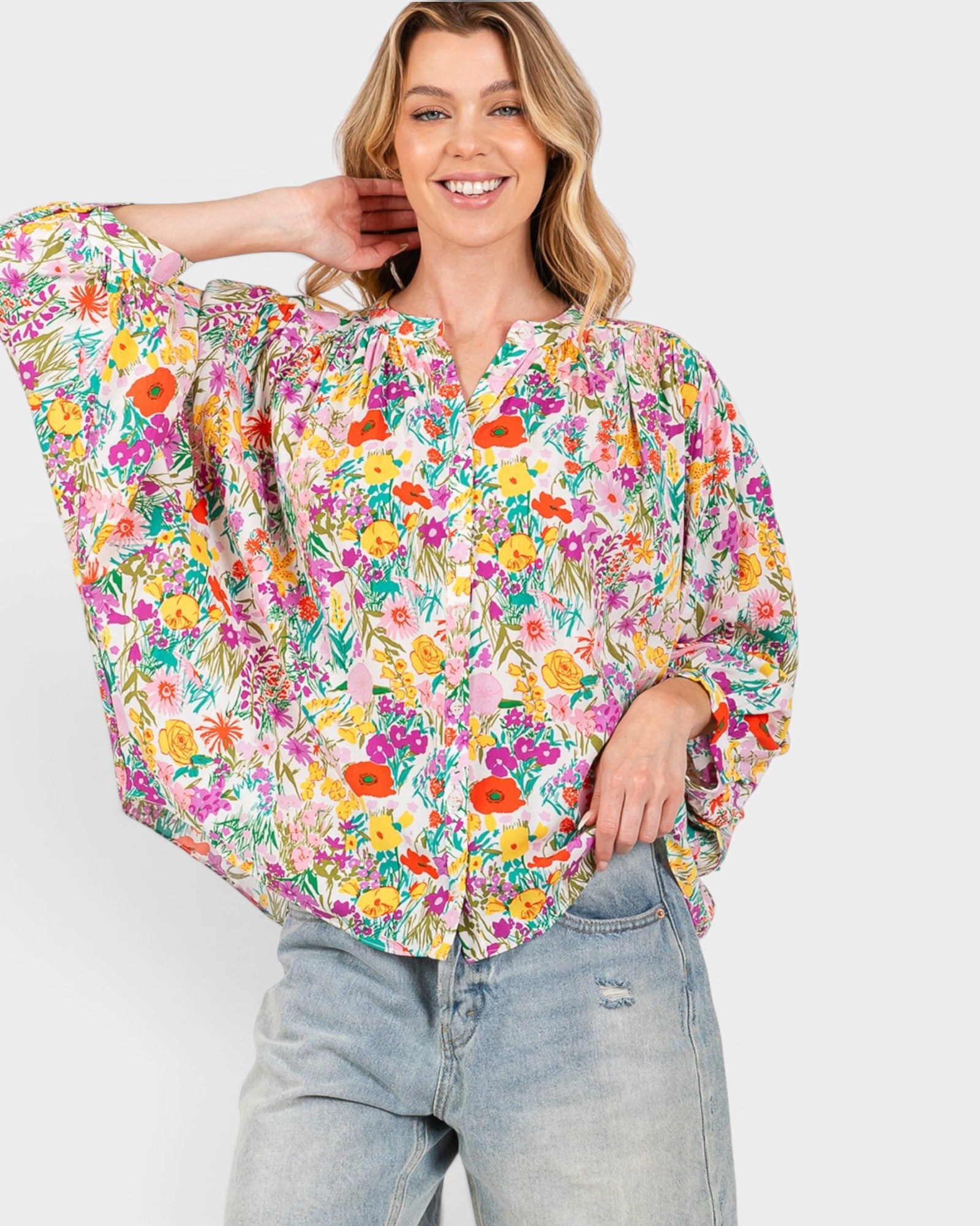 A woman wearing a vibrant, multicolor floral print blouse with balloon sleeves and light-wash distressed jeans, posing against a solid light gray background.