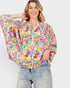 A woman wearing a vibrant, multicolor floral print blouse with balloon sleeves and light-wash distressed jeans, posing against a solid light gray background.
