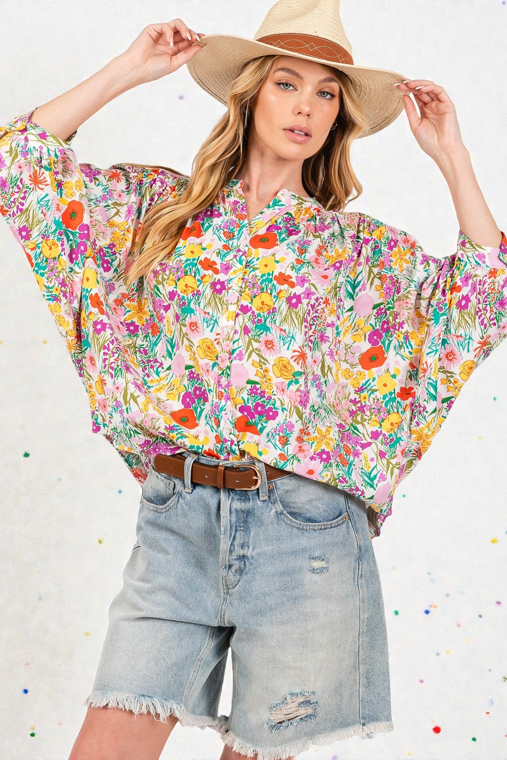 Woman wearing a colorful floral blouse and denim shorts with a wide-brimmed hat on a light background