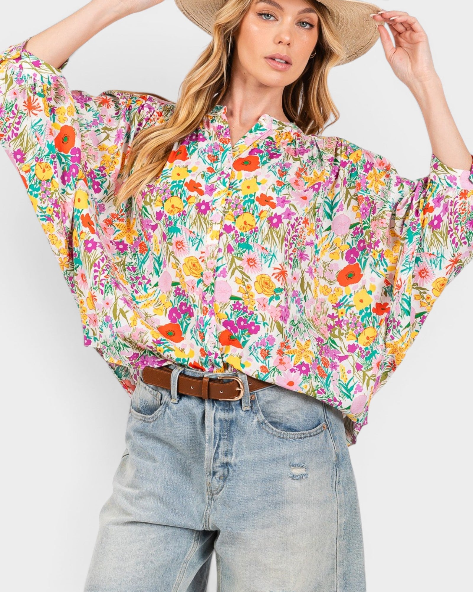 A woman wearing a colorful floral print blouse with wide sleeves, styled with light-wash jeans and a brown belt, against a plain light gray background.
