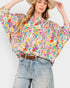 A woman wearing a colorful floral print blouse with wide sleeves, styled with light-wash jeans and a brown belt, against a plain light gray background.