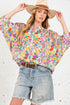 Woman wearing a colorful floral blouse and denim shorts with a wide-brimmed hat on a light background