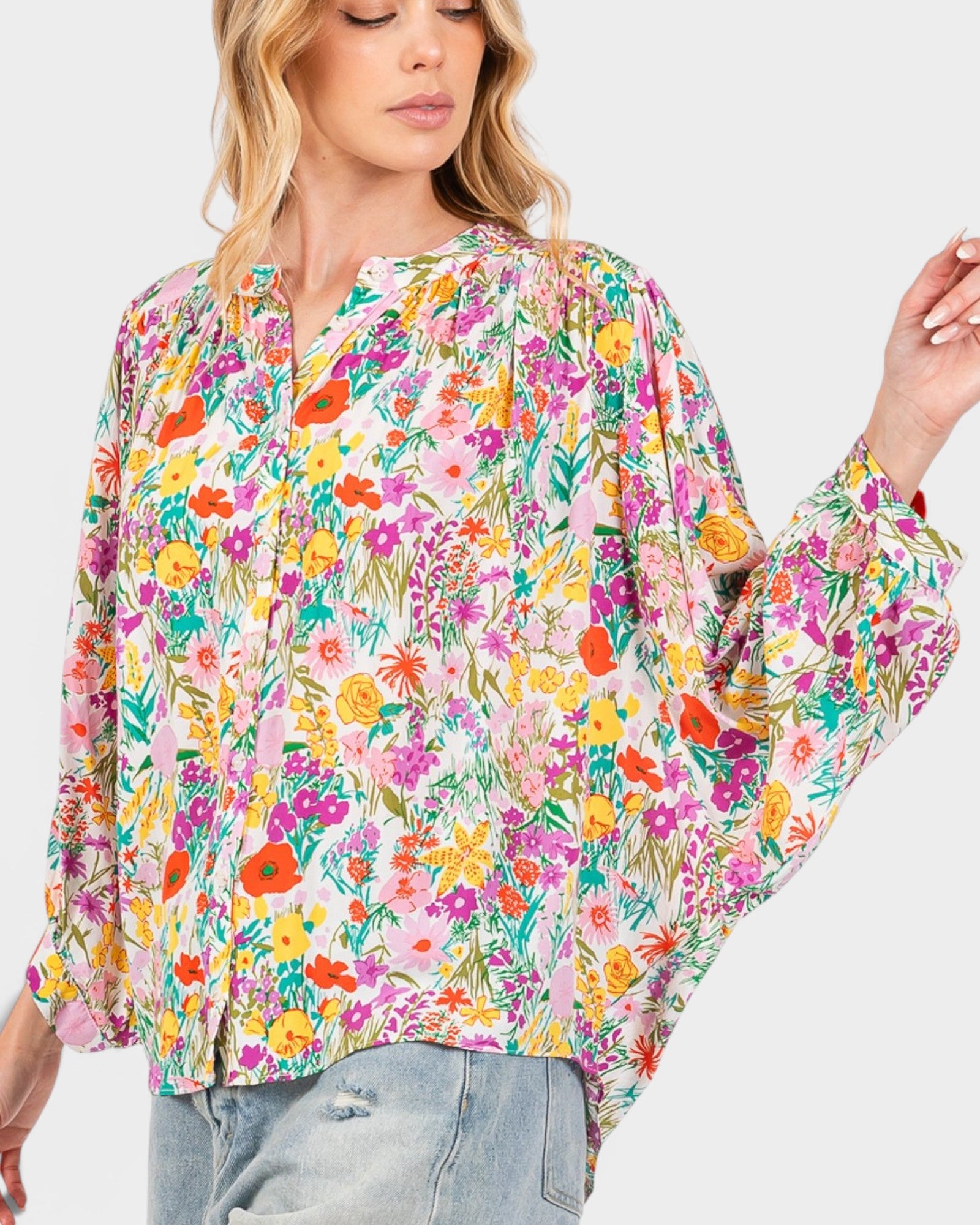 A woman wearing a long-sleeve blouse with a vibrant multicolor floral print on a light pink background, paired with light-wash jeans, against a solid light gray background.