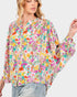 A woman wearing a long-sleeve blouse with a vibrant multicolor floral print on a light pink background, paired with light-wash jeans, against a solid light gray background.