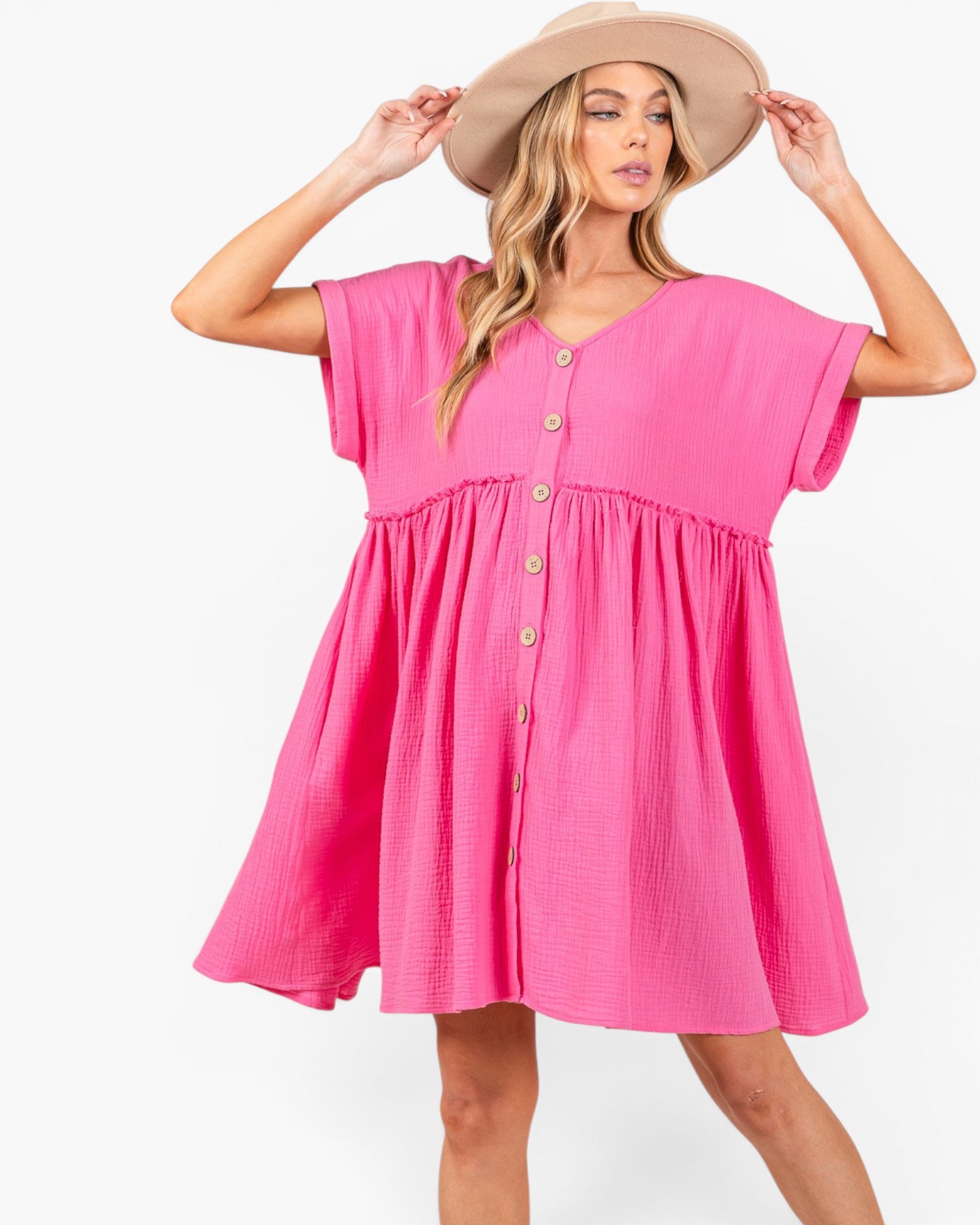 A woman wearing a bright pink short-sleeve babydoll dress with a button-down front and a beige wide-brim hat, posing against a plain white background.