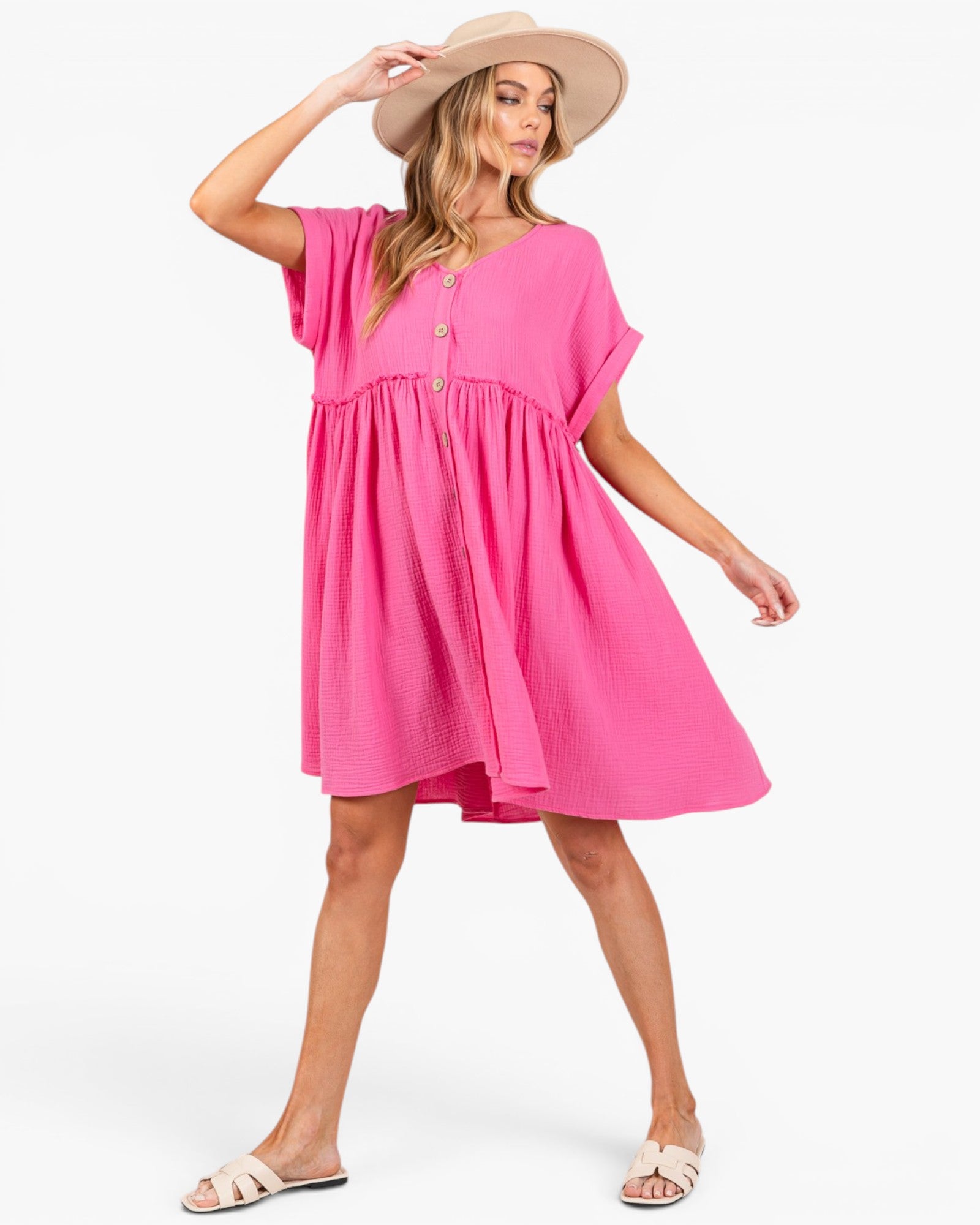 A woman modeling a bright pink short-sleeve babydoll dress with a buttoned neckline, wearing a beige wide-brim hat and beige slide sandals against a plain white background.