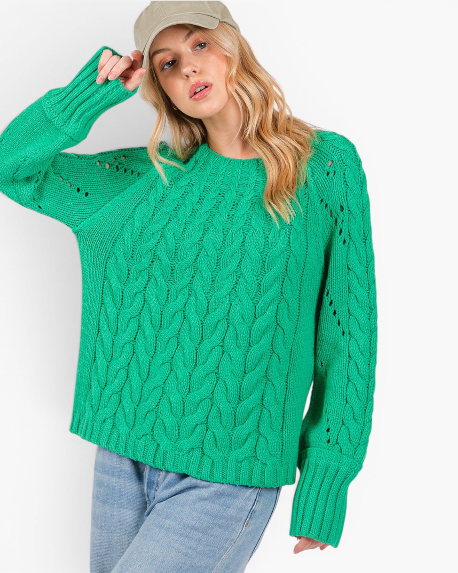 A woman wearing a bright green cable-knit sweater, light blue jeans, and a beige baseball cap, posing against a plain white studio background.