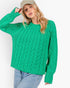A woman wearing a bright green cable-knit sweater, light blue jeans, and a beige baseball cap, posing against a plain white studio background.