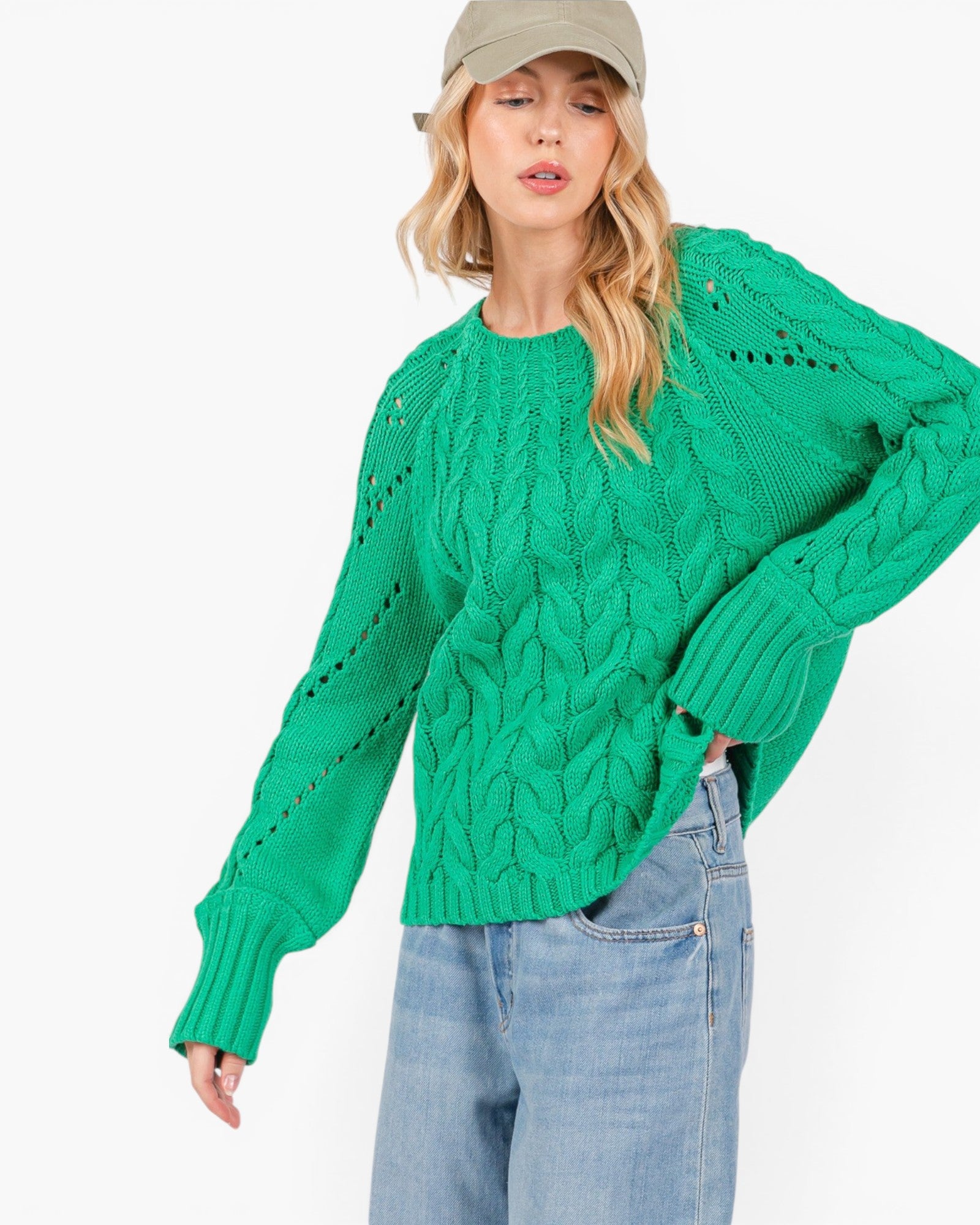A woman wearing a bright green cable-knit sweater, light-wash blue jeans, and a beige baseball cap, photographed against a plain white studio background.