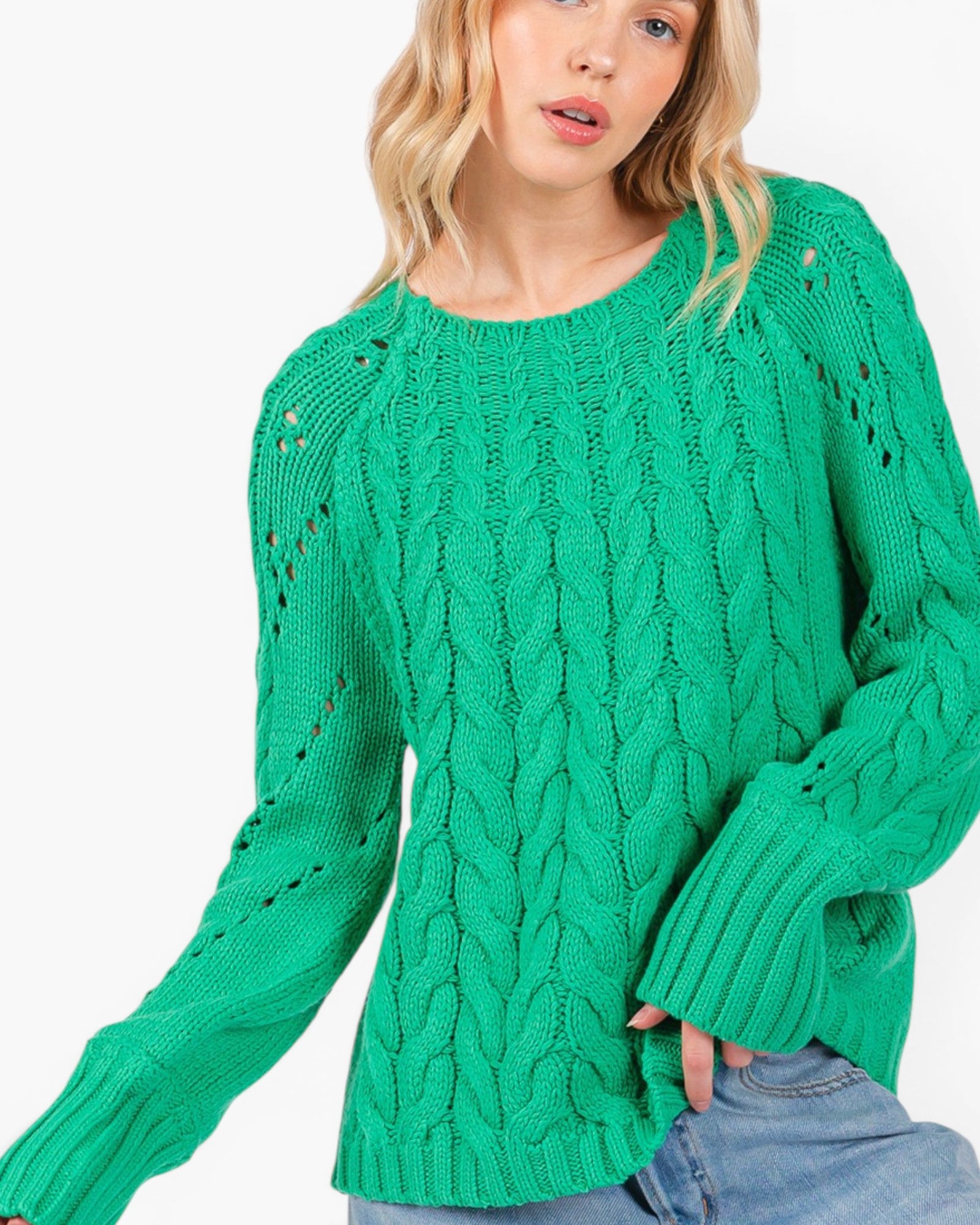 A woman wearing a bright green cable-knit sweater with textured knit details and wide sleeves, paired with blue jeans, against a plain white background.
