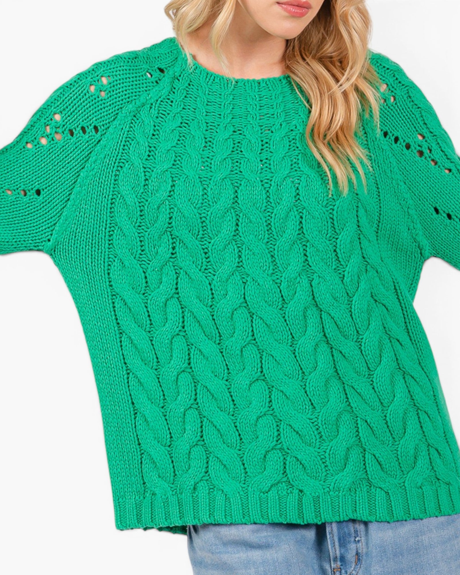 A woman wearing a bright green cable-knit sweater with textured knit details and eyelet cutouts on the sleeves, paired with blue jeans against a plain white background.