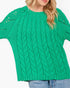 A woman wearing a bright green cable-knit sweater with textured knit details and eyelet cutouts on the sleeves, paired with blue jeans against a plain white background.