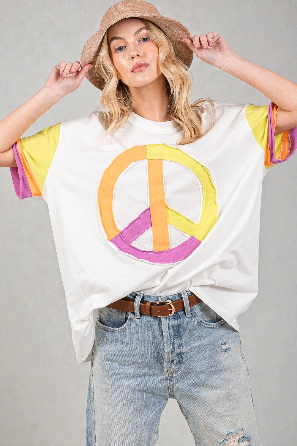 Person wearing a white t-shirt with a colorful peace symbol design, light blue jeans, and a straw hat on a gray background.