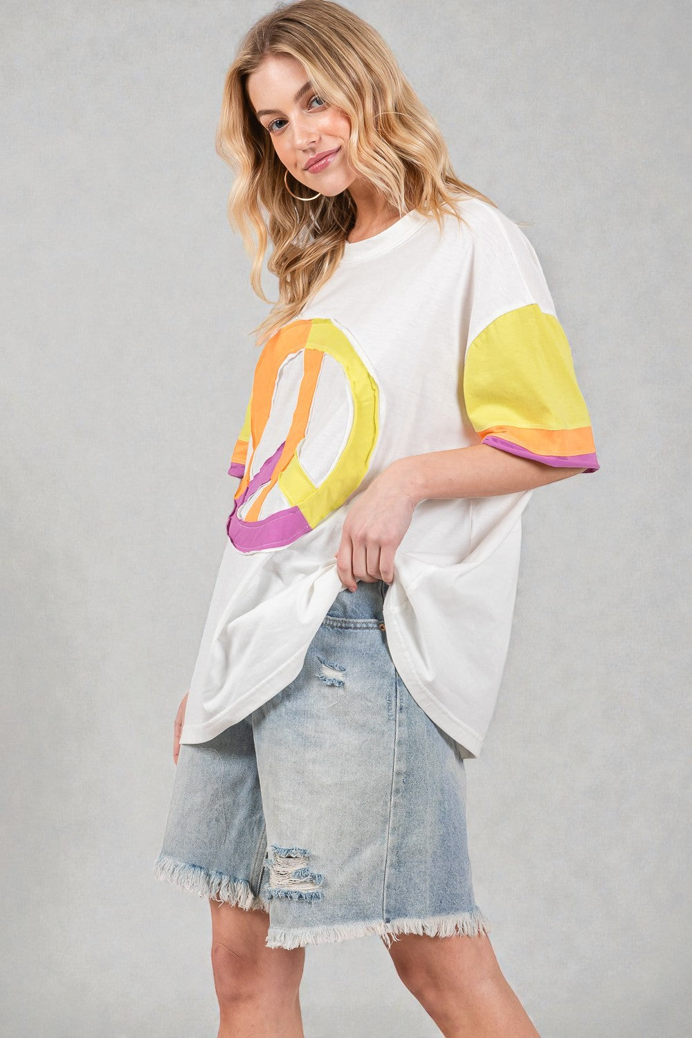 Woman wearing a white t-shirt with a colorful peace symbol design and denim shorts on a gray background