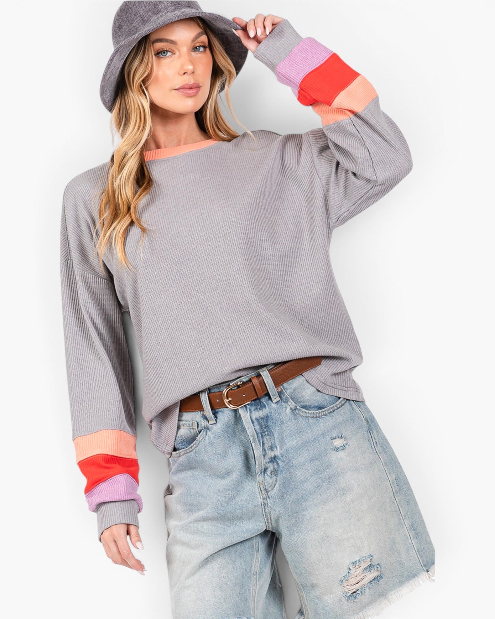 A woman modeling a gray long-sleeve knit sweater with colorful striped cuffs in pink, red, and orange, paired with light-wash distressed denim shorts, a brown belt, and a gray bucket hat against a white studio background.