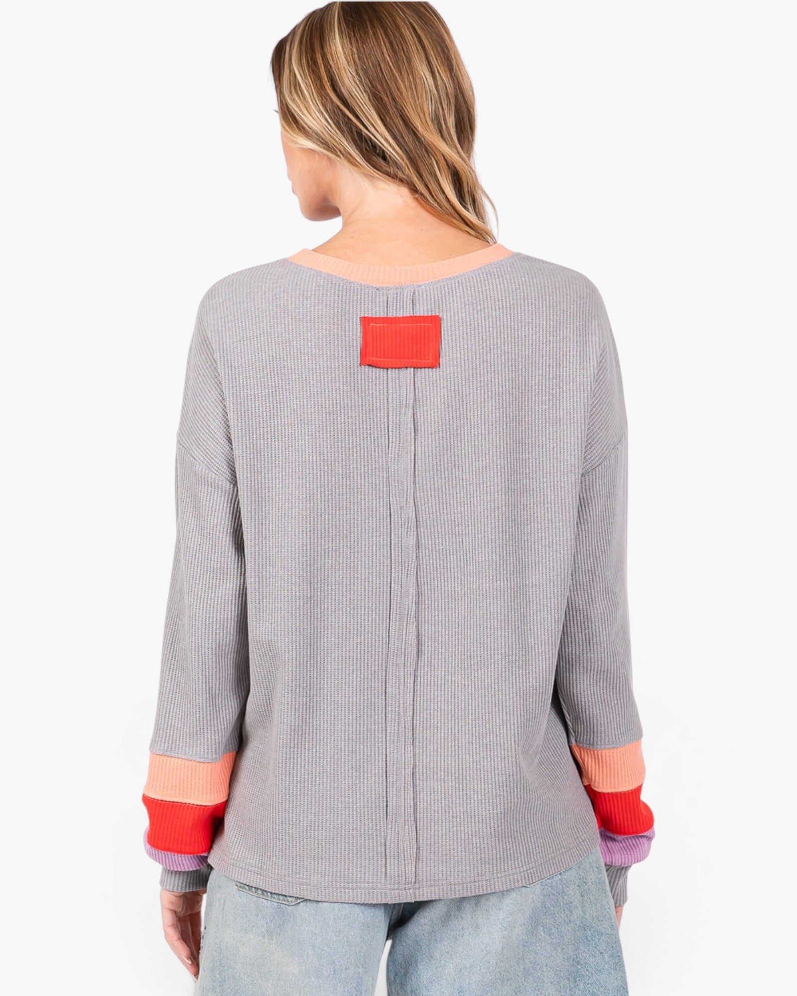 A woman shown from the back wearing a gray ribbed knit long-sleeve top with a red rectangular patch at the back neckline and red and orange striped cuffs, paired with light blue jeans against a plain white background.