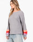 A woman wearing a gray waffle-knit long-sleeve top with colorful striped cuffs in orange, pink, and purple, paired with light-wash jeans, against a plain white background.