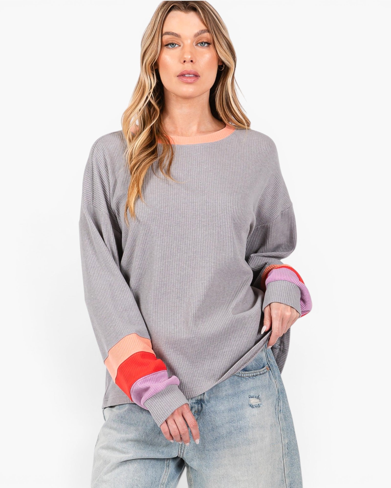 A woman modeling a gray long-sleeve knit sweater with color-blocked cuffs in red, pink, and purple, paired with light-wash jeans against a plain white studio background.