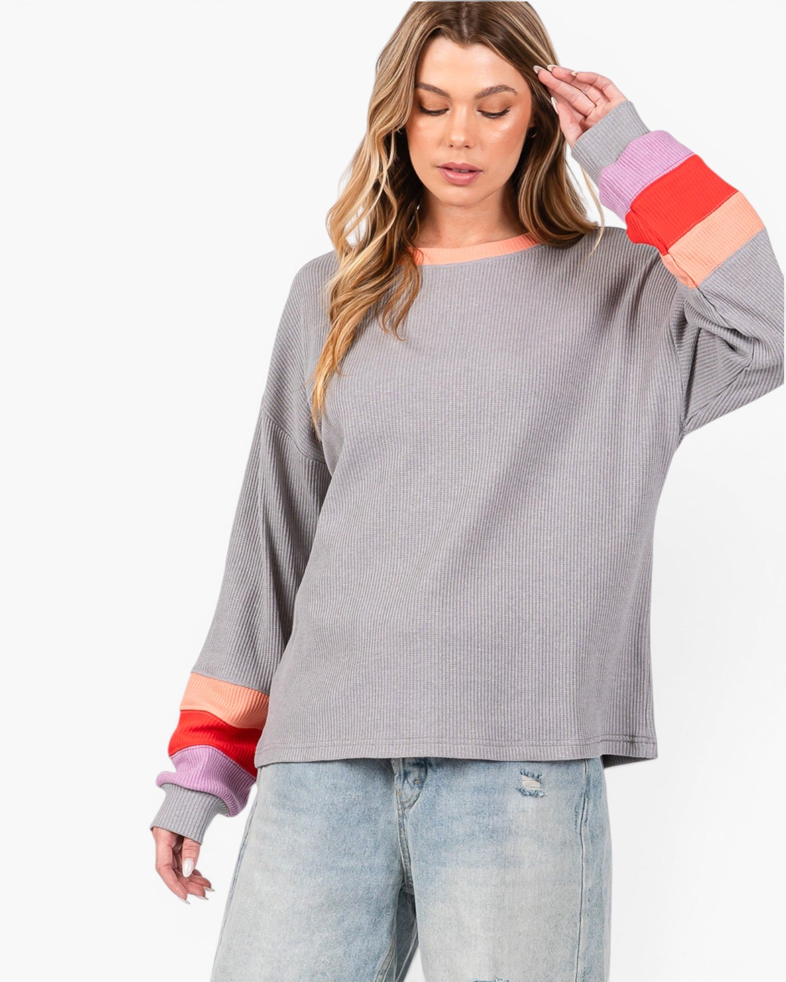 A woman wearing a gray long-sleeve ribbed knit top with colorful striped cuffs in pink, orange, and red, paired with light blue jeans, against a plain white background.