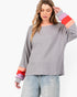 A woman wearing a gray long-sleeve ribbed knit top with colorful striped cuffs in pink, orange, and red, paired with light blue jeans, against a plain white background.