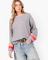 A woman modeling a gray long-sleeve knit sweater with colorful striped cuffs in pink, orange, and red, paired with light-wash jeans and a brown belt against a plain white background.