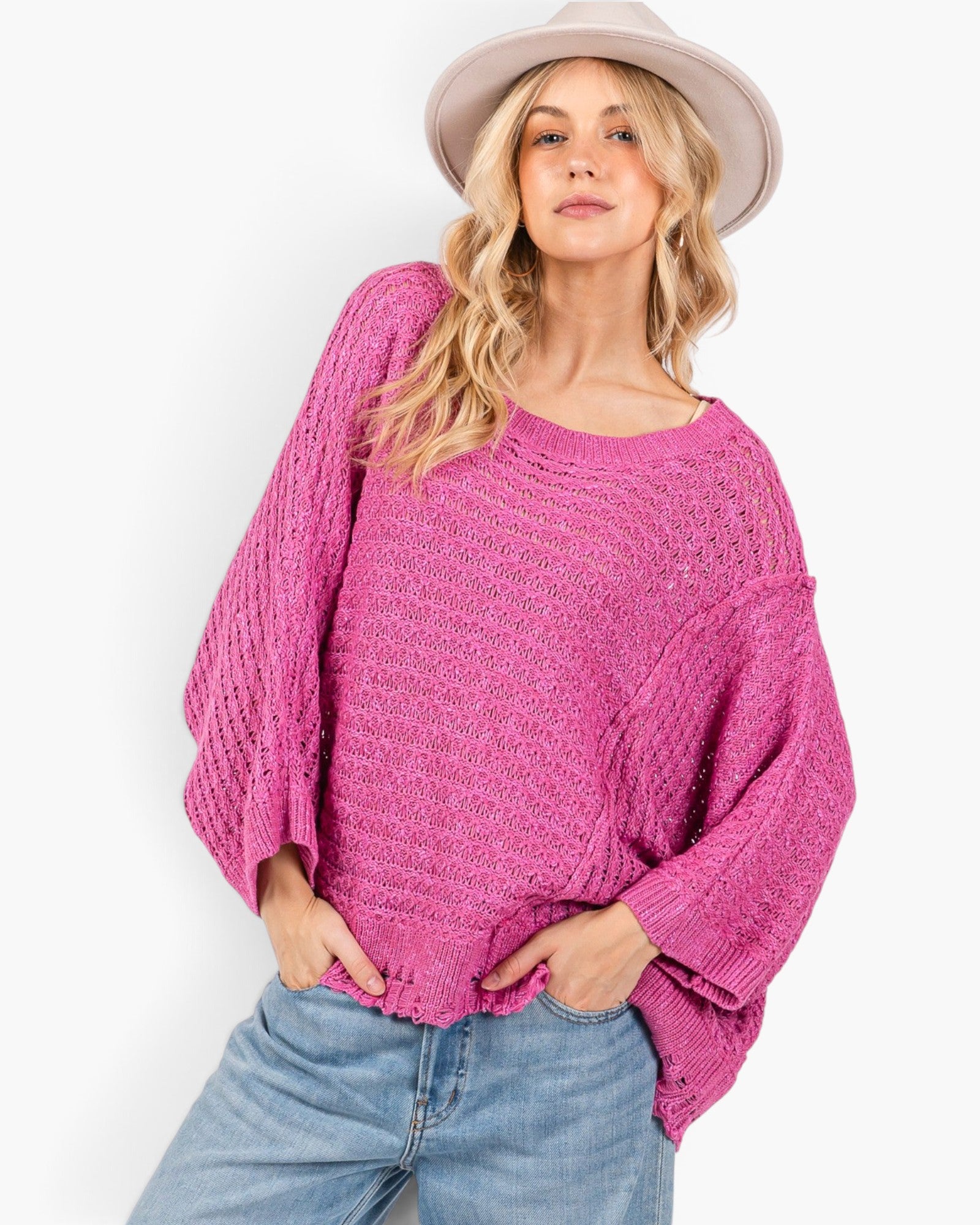 A woman wearing a bright pink open-knit sweater with balloon sleeves, paired with light blue jeans and a beige wide-brim hat, against a plain white background.