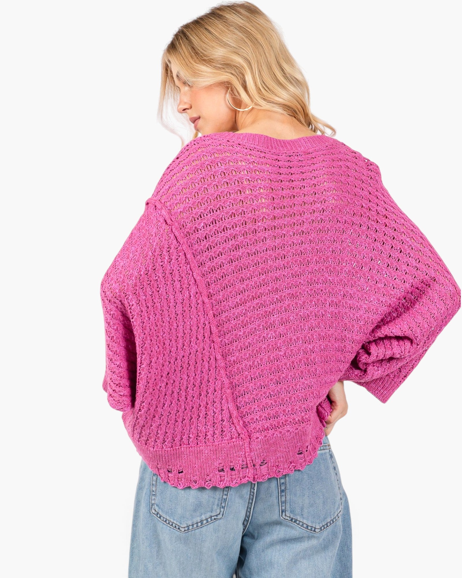A woman shown from the back wearing a bright pink chunky knit sweater and light blue jeans against a plain white background.