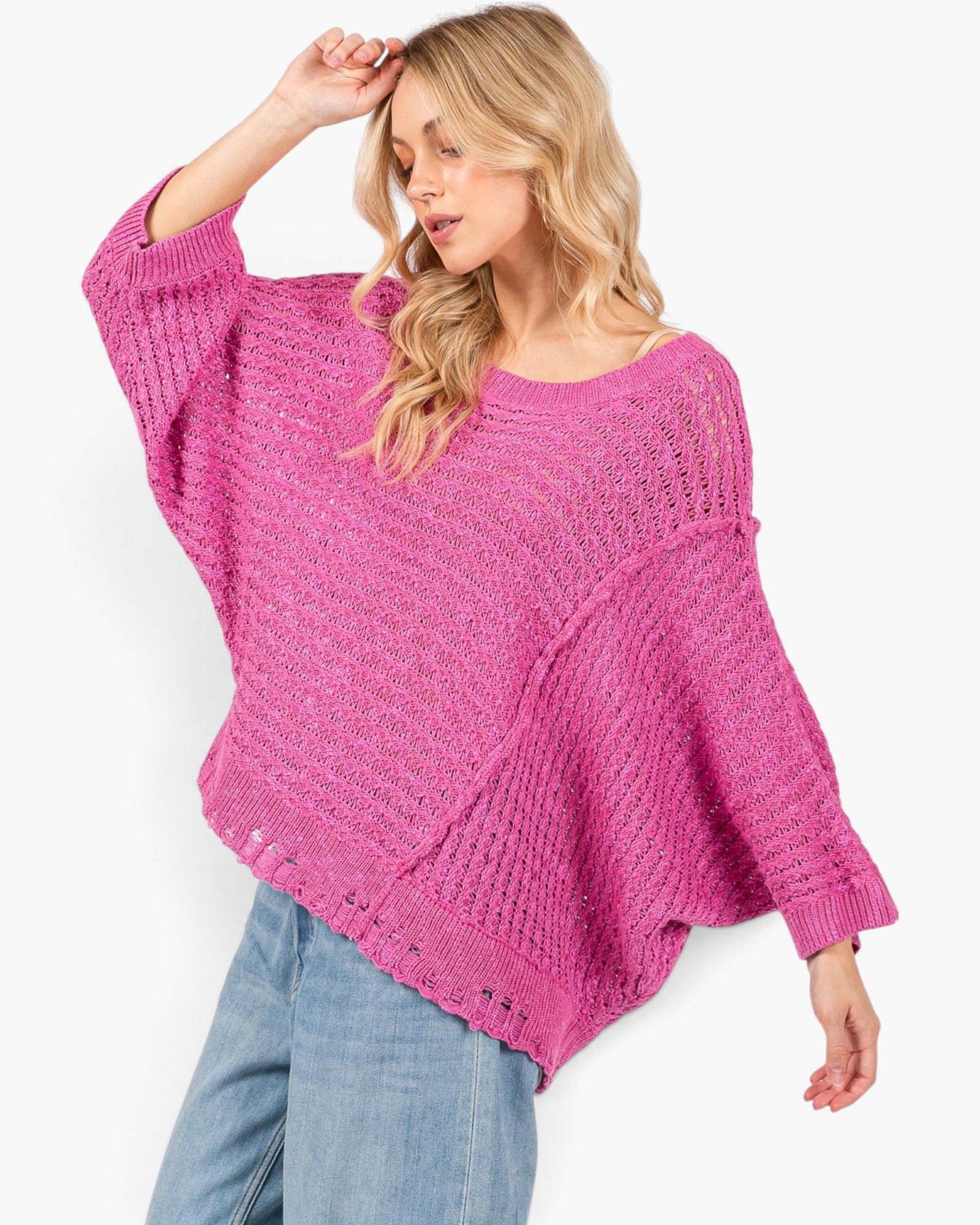 A woman wearing a bright pink open-knit sweater with wide batwing sleeves and light blue jeans, posed against a clean white background.