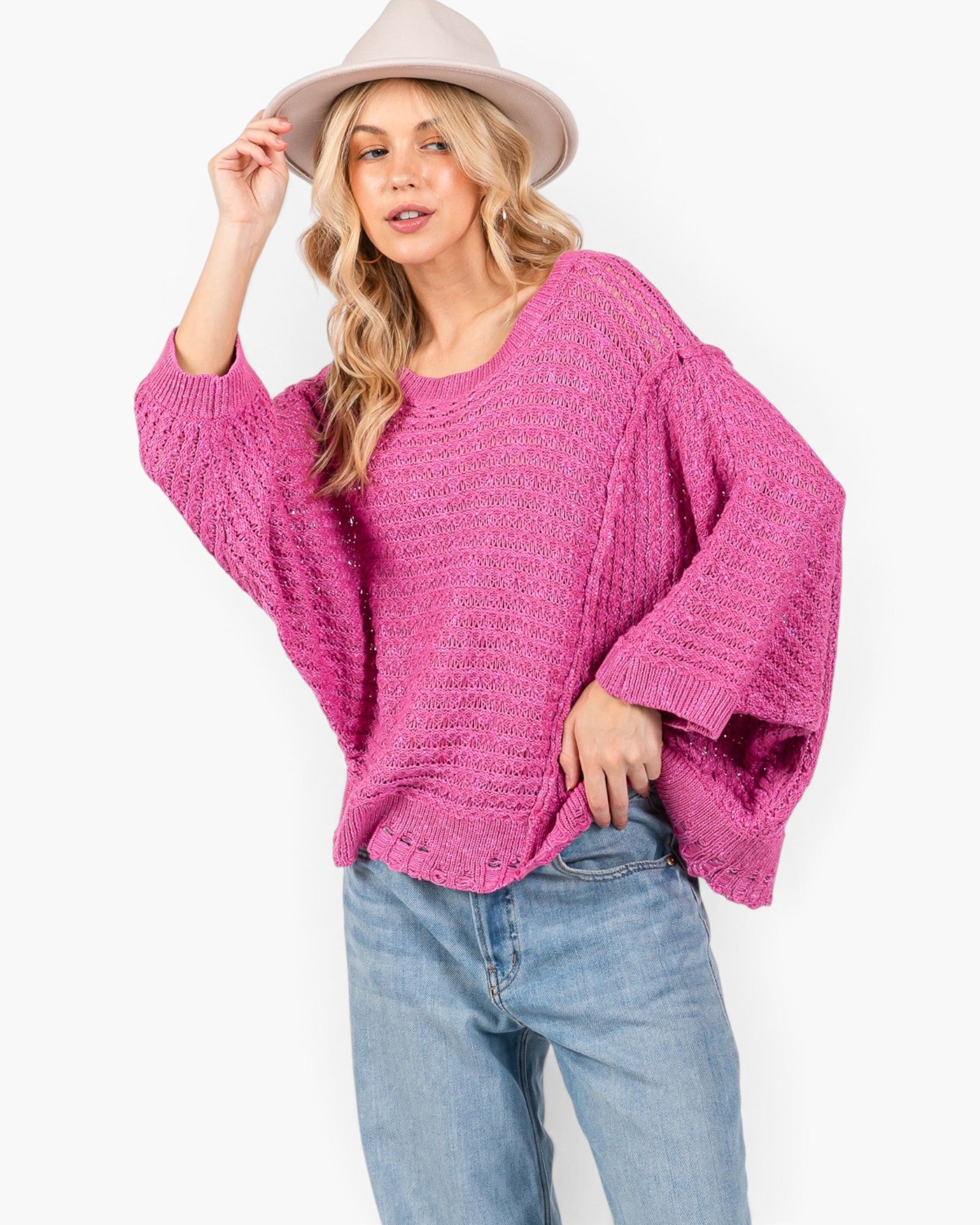 A woman wearing a chunky knit magenta pink sweater with wide draped sleeves, paired with light blue jeans and a beige wide-brim hat, against a plain white background.
