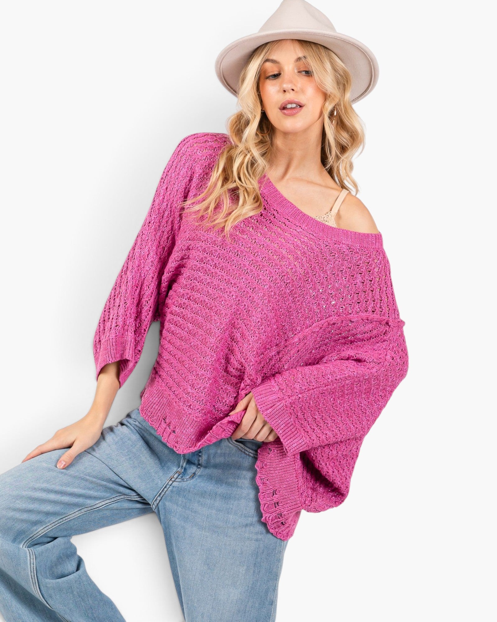 A woman wearing a bright pink chunky knit off-the-shoulder sweater with bell sleeves, paired with light blue jeans and a beige wide-brim hat, posing against a white studio background.