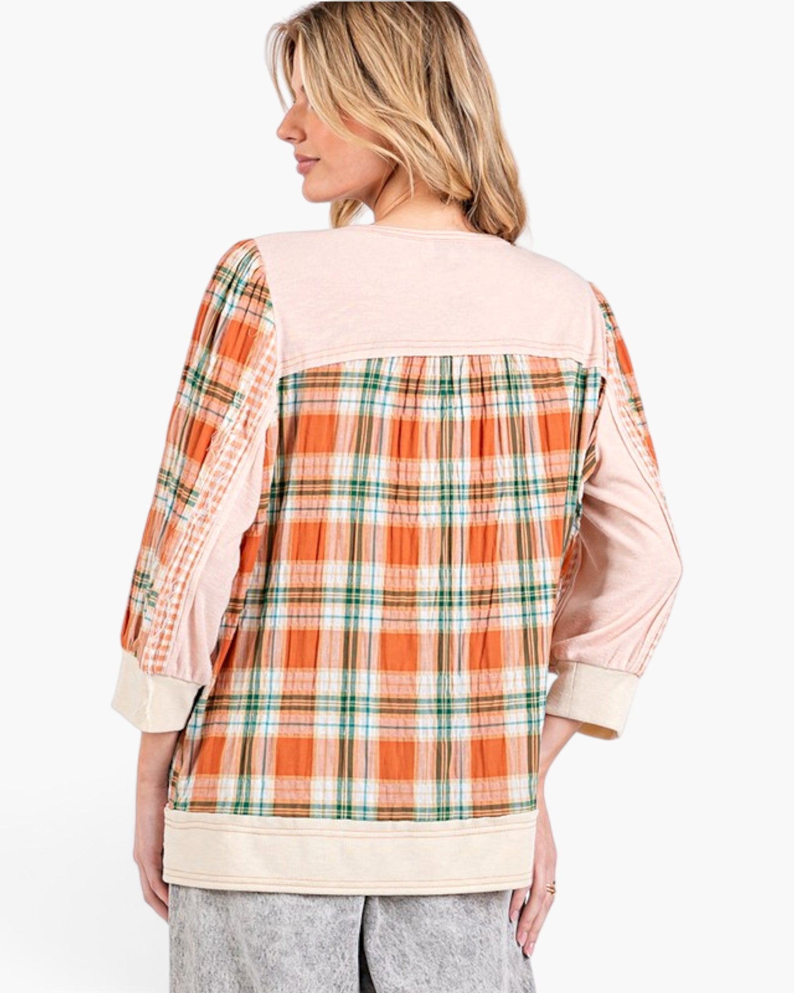 A woman shown from the back wearing a patchwork-style plaid top with orange, green, and white plaid panels, light pink sleeves and yoke, and cream cuffs and hem, paired with gray pants against a plain white studio background.