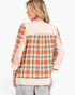 A woman shown from the back wearing a patchwork-style plaid top with orange, green, and white plaid panels, light pink sleeves and yoke, and cream cuffs and hem, paired with gray pants against a plain white studio background.