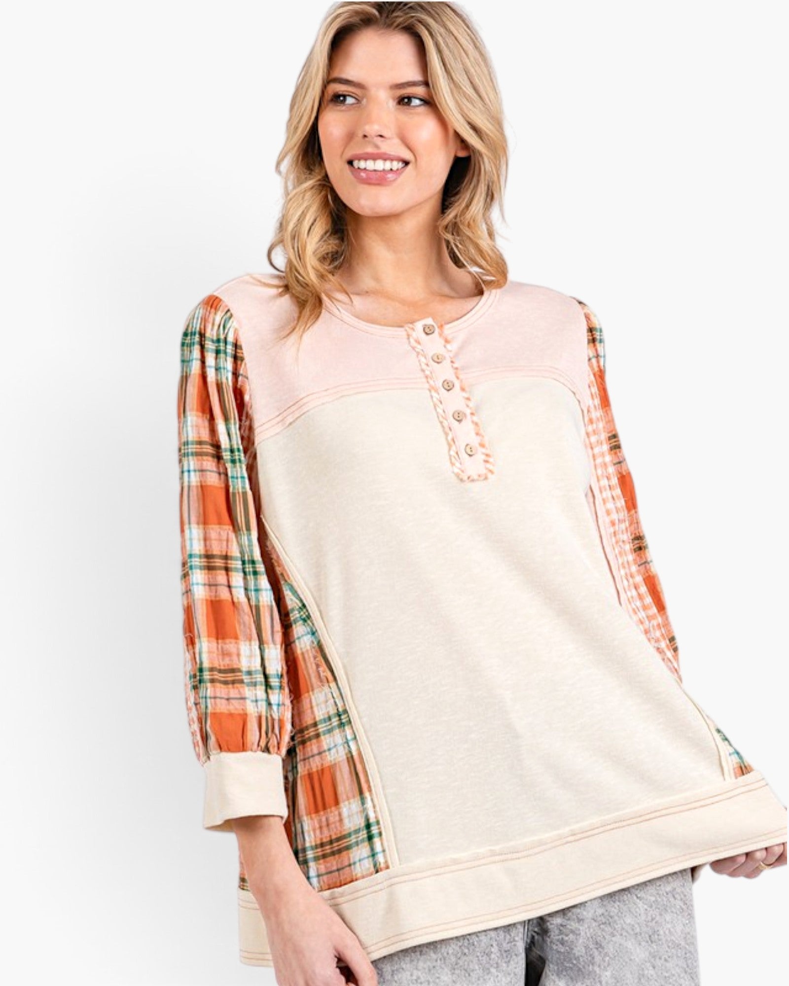 A woman modeling a color-block knit top with a cream body, a light pink henley neckline, and orange plaid sleeves, set against a plain white studio background.