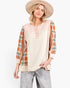 A woman modeling a color-blocked long-sleeve top with a cream body, peach yoke, and orange plaid sleeves, paired with gray jeans and a beige wide-brim hat against a white studio background.