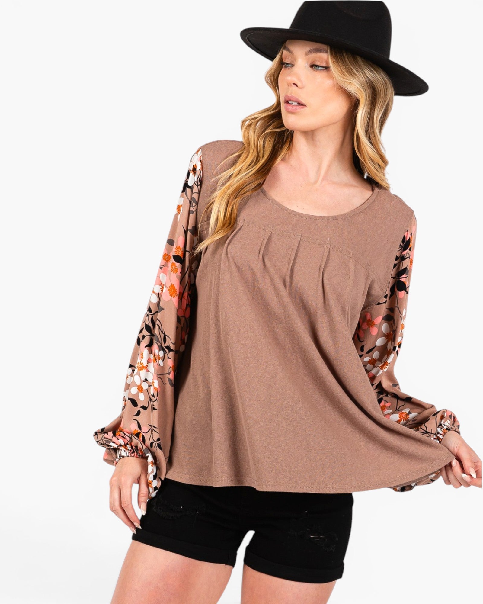 A woman wearing a brown scoop-neck top with floral printed long sleeves, paired with black shorts and a black wide-brim hat, against a plain white background.