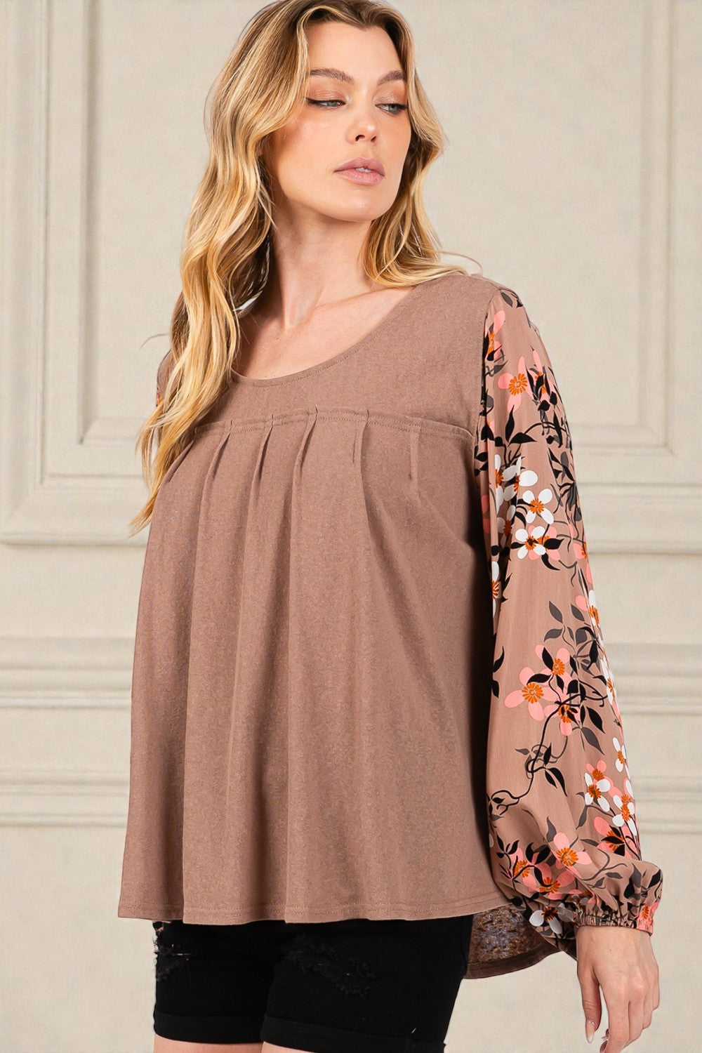 Woman wearing a brown top with floral sleeves against a beige wall.