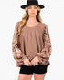 A woman modeling a brown long-sleeve top with a floral pattern on the sleeves, paired with black shorts and a black hat, against a plain white background.