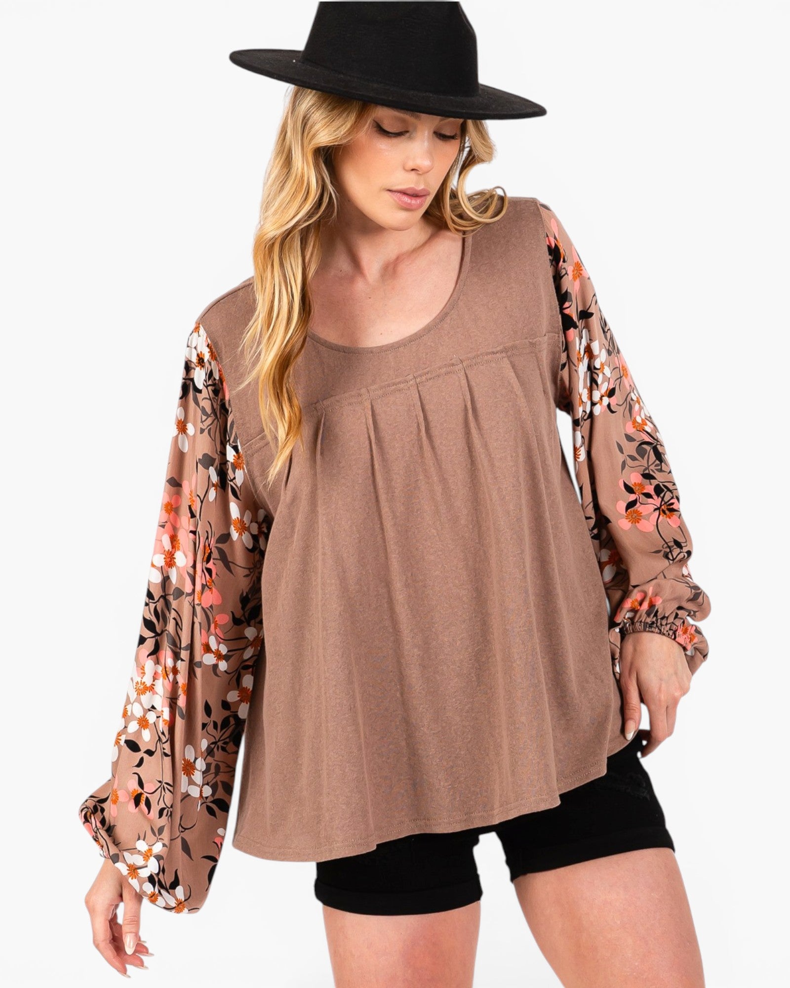 A woman wearing a brown scoop-neck top with long sheer floral sleeves, paired with black shorts and a black hat, against a plain white background.