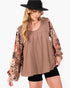 A woman wearing a brown scoop-neck top with long sheer floral sleeves, paired with black shorts and a black hat, against a plain white background.