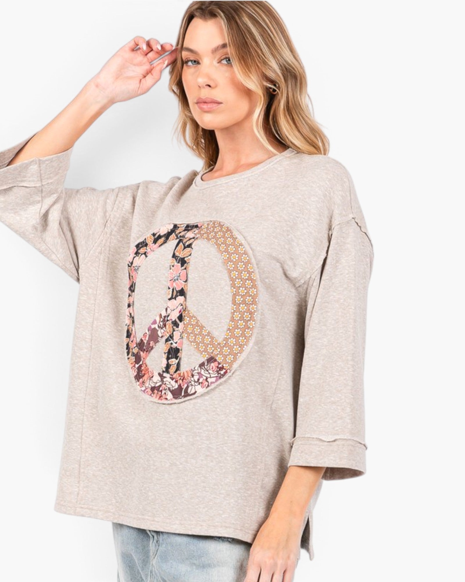 A woman wearing a light beige heathered oversized sweatshirt with a large peace sign graphic on the front, featuring a floral pattern and a geometric pattern, paired with light blue jeans against a plain white background.