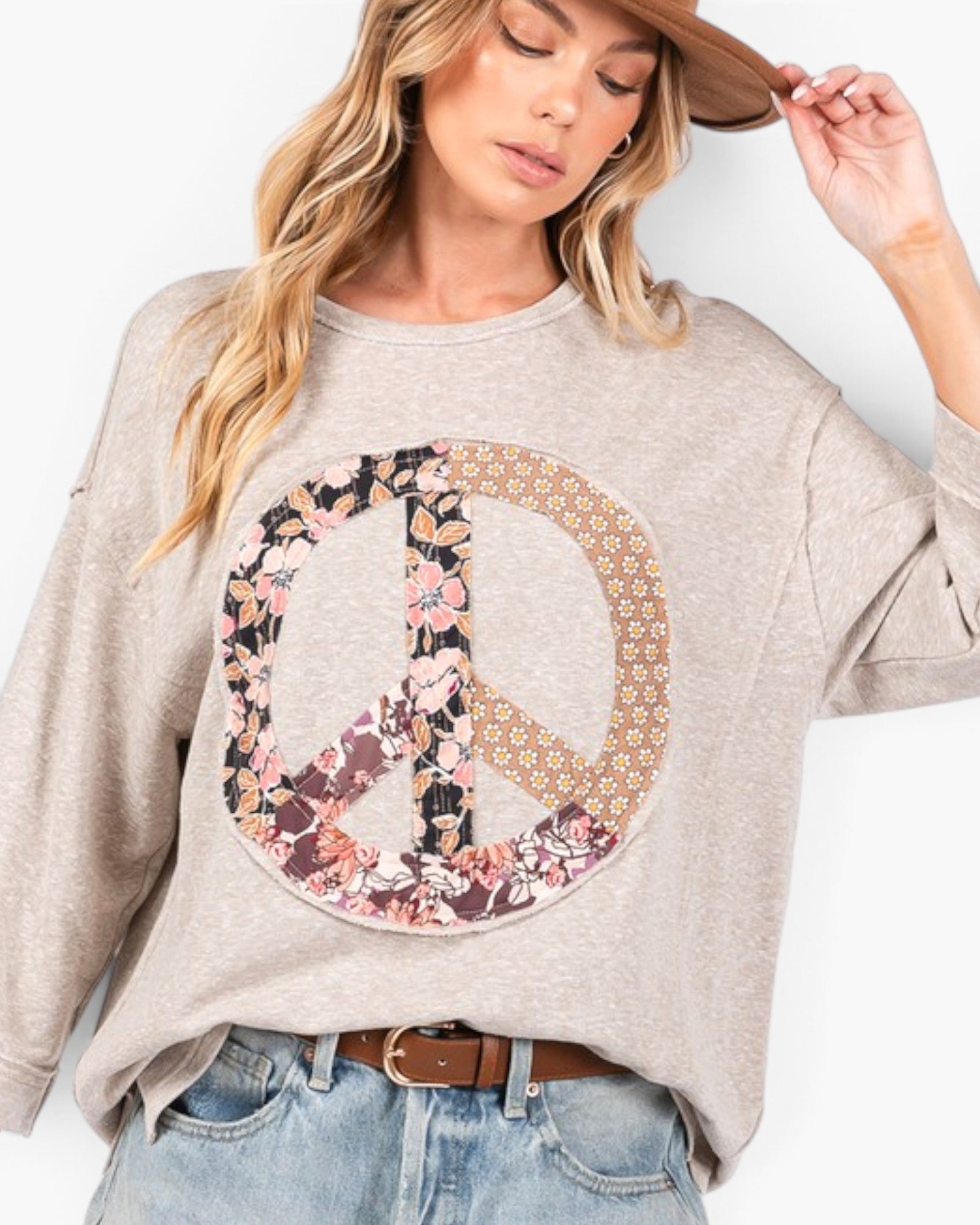 A woman wearing a light heather gray long-sleeve sweatshirt with a large peace sign graphic on the front, featuring a floral pattern on one side and a tan geometric pattern on the other. She is also wearing blue jeans, a brown belt, and a brown hat against a plain white background.