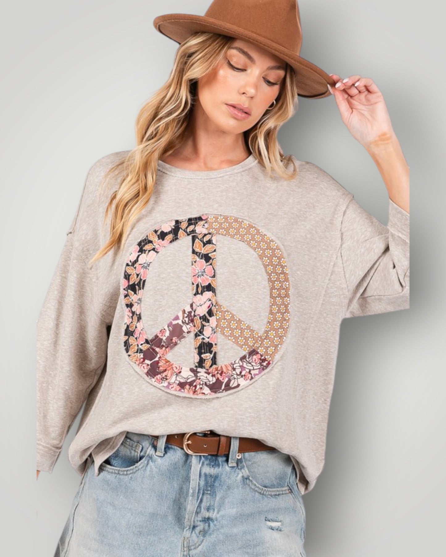 Woman wearing a beige sweatshirt with a floral peace symbol design, light blue jeans, and a brown hat.