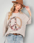 Woman wearing a beige sweatshirt with a floral peace symbol design, light blue jeans, and a brown hat.