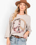 A woman wearing a heather taupe crewneck sweatshirt with a large peace sign graphic filled with floral and paisley patterns, styled with light-wash jeans, a brown belt, and a brown wide-brim hat against a white studio background.