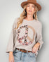 Woman wearing a beige sweater with a floral peace sign design, blue jeans, and a brown hat on a plain background.