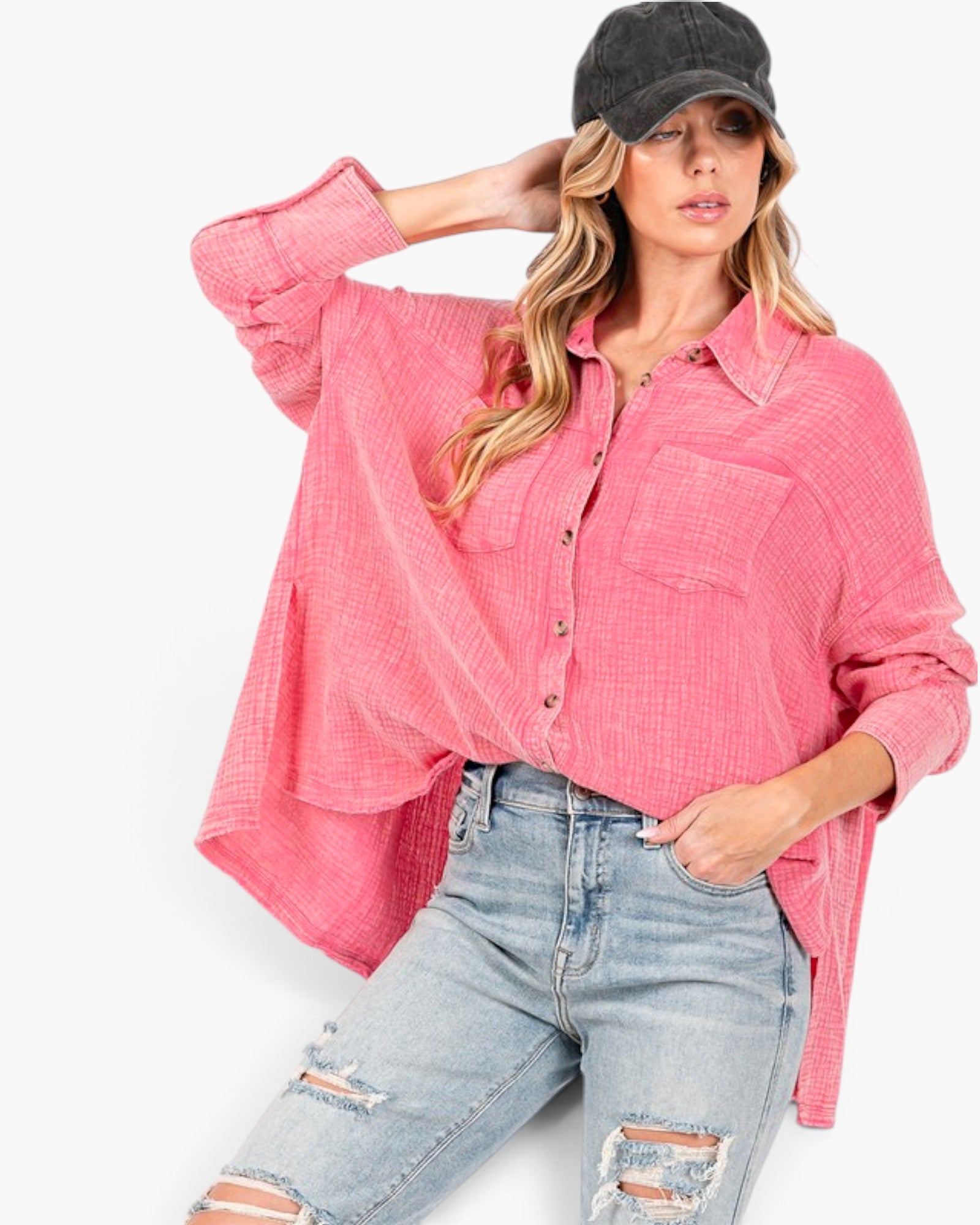 A woman wearing a pink textured button-up shirt with light-wash distressed jeans and a black baseball cap, posed against a plain white studio background.