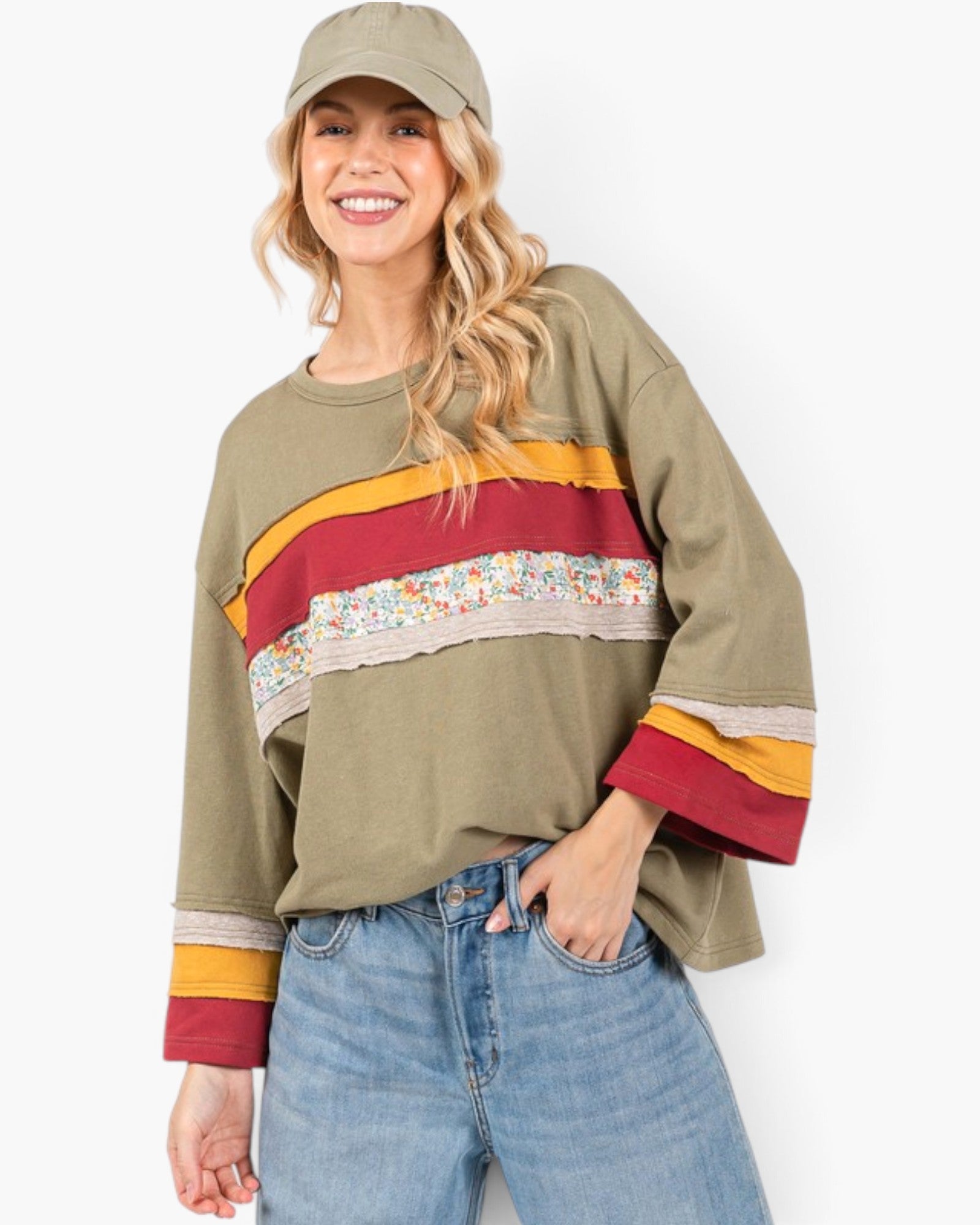 A woman wearing a cropped olive green sweatshirt with colorful horizontal stripes across the chest and cuffs, paired with blue jeans and a matching olive green cap, against a plain white background.