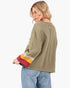 A woman shown from the back wearing an olive green cropped sweatshirt with colorful striped cuffs in red, yellow, and beige, paired with light blue jeans, against a plain white background.